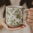 Load image into Gallery viewer, A ceramic mug with a green botanical floral pattern on a cream background, held in two hands. The mug features a solid beige glazed base at the bottom and a white handle.