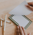Load image into Gallery viewer, A close-up of a hand holding a sheet of white paper over a lined notepad with green striped borders, placed on a light wood desk with two pens nearby.