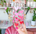 Load image into Gallery viewer, A hand with pink nail polish holds a clear glass can-shaped tumbler decorated with layered heart stickers in shades of red, pink, and light lavender, set against a softly blurred background of shelves and plants.