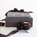 Load image into Gallery viewer, A dark brown candle in an amber glass jar with its lid off, styled on top of a dark brown gift box with a brown satin ribbon and the board game 'CLUE'.
