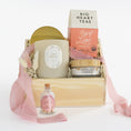 Load image into Gallery viewer, A curated gift basket set in a light wood crate with shredded paper filler, featuring a large candle, a tea box, a small jar, and a pink ribbon, photographed on a clean white background.