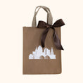 Load image into Gallery viewer, A reusable brown gift tote with beige handles, decorated with a white Kansas City skyline graphic and a large dark brown satin bow, set against a solid cream background.
