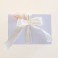 Load image into Gallery viewer, A square white gift box wrapped with a large cream satin ribbon bow and tied with dried pastel grass stems, photographed on a warm off-white background.