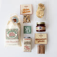 Load image into Gallery viewer, A flat-lay arrangement of Louisburg Ciderworks gift items on a white background, including a large bag of cider donut mix, a jar of apple cider, cinnamon sticks, apple slices, spices, and a tin of spiced apple cider.