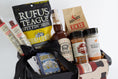 Load image into Gallery viewer, A barbecue gift basket in a black and yellow utility tote, a bag of Rufus Teague Spittin' Seeds sunflowers, a bottle of BBQ Sauce, a jars of spices and seasonings, and a patterned towel with barbecue bottle illustrations, photographed on a white background.