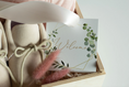 Load image into Gallery viewer, A close-up of a gift box containing cream-colored baby booties tied with beige rope, a white welcome card with green botanical illustrations and gold script, and pink dried grass stems on a light background.