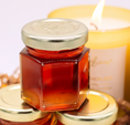 Load image into Gallery viewer, A hexagonal glass jar filled with amber-red honey, topped with a silver metal lid, displayed on top of other similar jars with a softly blurred yellow candle in the background.