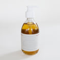 Load image into Gallery viewer, A clear glass pump bottle filled with amber-colored hand soap, featuring a white pump dispenser and a white label with fine blue and red text, set against a plain white background.