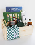 Load image into Gallery viewer, A wooden gift basket filled with golf-themed items, including a book titled 'Remarkable Golf Courses' by Iain Spragg, a green textured pillow with multi-colored golf tees, a green checkered towels, a white candle with a green plaid wax se
