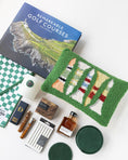 Load image into Gallery viewer, A curated flat lay of golf-themed merchandise on a white background, including a book titled 'Remarkable Golf Courses' by Iain Spragg, a green textured pillow with multi-colored golf tees, a green checkered towels, a white candle with a green plaid wax seal, bourbon-soaked toothpicks in both glass jars and cardboard packaging, a shoe shine kit in a wood grain box, silver plated cocktail picks, a bottle of black walnut cocktail bitters, and a set of green leather coasters.