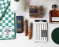 Load image into Gallery viewer, A curated flat lay of golf-themed merchandise on a white background, including a book titled 'Remarkable Golf Courses' by Iain Spragg, a green textured pillow with multi-colored golf tees, a green checkered towels, a white candle with a green plaid wax seal, bourbon-soaked toothpicks in both glass jars and cardboard packaging, a shoe shine kit in a wood grain box, silver plated cocktail picks, a bottle of black walnut cocktail bitters, and a set of green leather coasters.