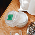 Load image into Gallery viewer, A clear ribbed glass jar candle with its lid off, showing white wax inside. The lid has a green square label with a black botanical-style logo.