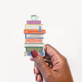 Load image into Gallery viewer, A hand with dark skin and burgundy nail polish holds a die-cut sticker featuring a colorful stack of books topped with a small coffee cup, against a plain white background.