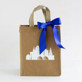 Load image into Gallery viewer, A brown kraft paper gift bag with beige handles, decorated with a white silhouette of the Kansas City skyline and a large royal blue satin bow, set against a plain white background.