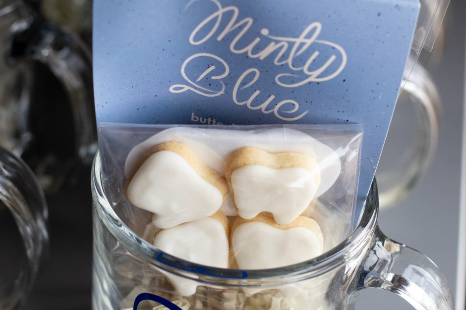 Teeth-shaped sugar cookies with white icing, packaged in a clear bag with a blue pack of tea labeled 'Minty Blue' and displayed in a glass mug.