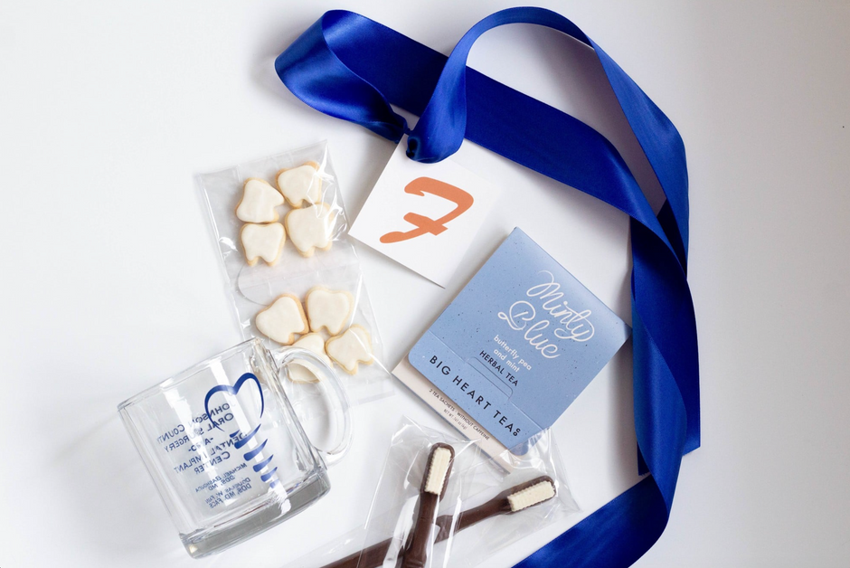 A flat-lay of a custom gift set on a white background, featuring a blue ribbon, a glass mug with blue graphics, teeth-shaped cookies, a blue tea box, and a small card with the client logo.