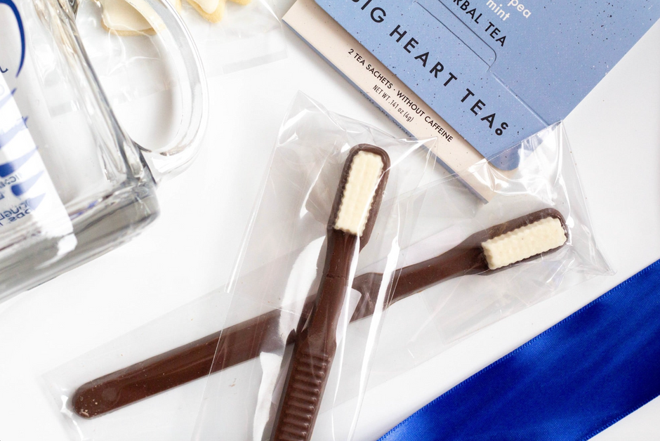 Chocolate toothbrushes in clear plastic packaging on a white surface, with a blue tea packet and other small items nearby.