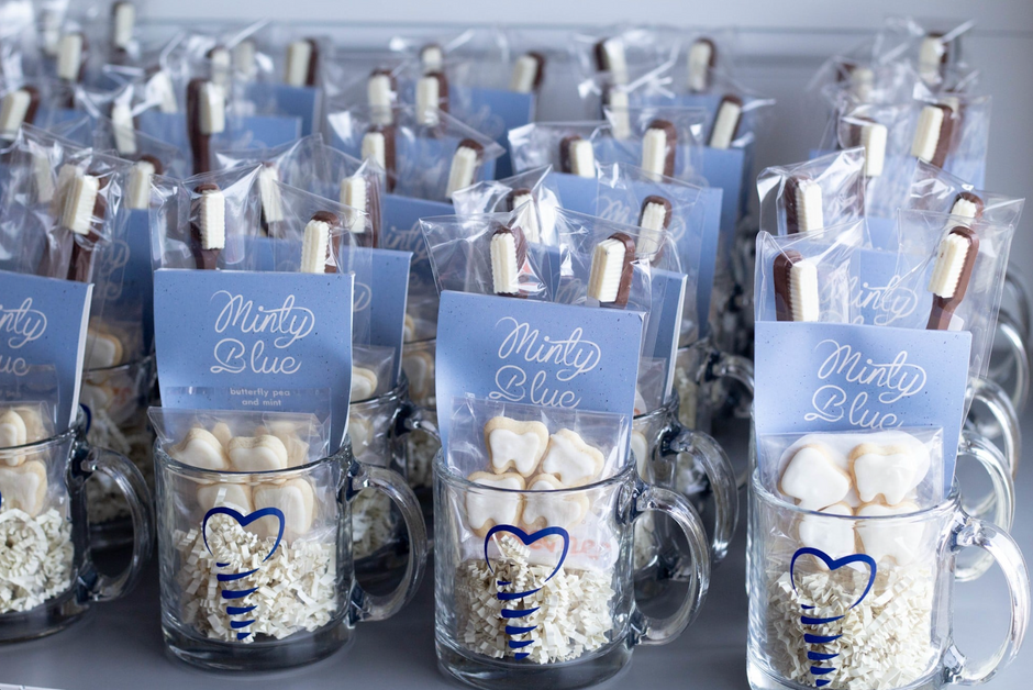 Several packaged chocolate toothbrush treats arranged in clear glass mugs on a shelf. Each package also includes a blue pack of tea reading 'Minty Blue,' and small teeth-shaped cookies in clear packaging.