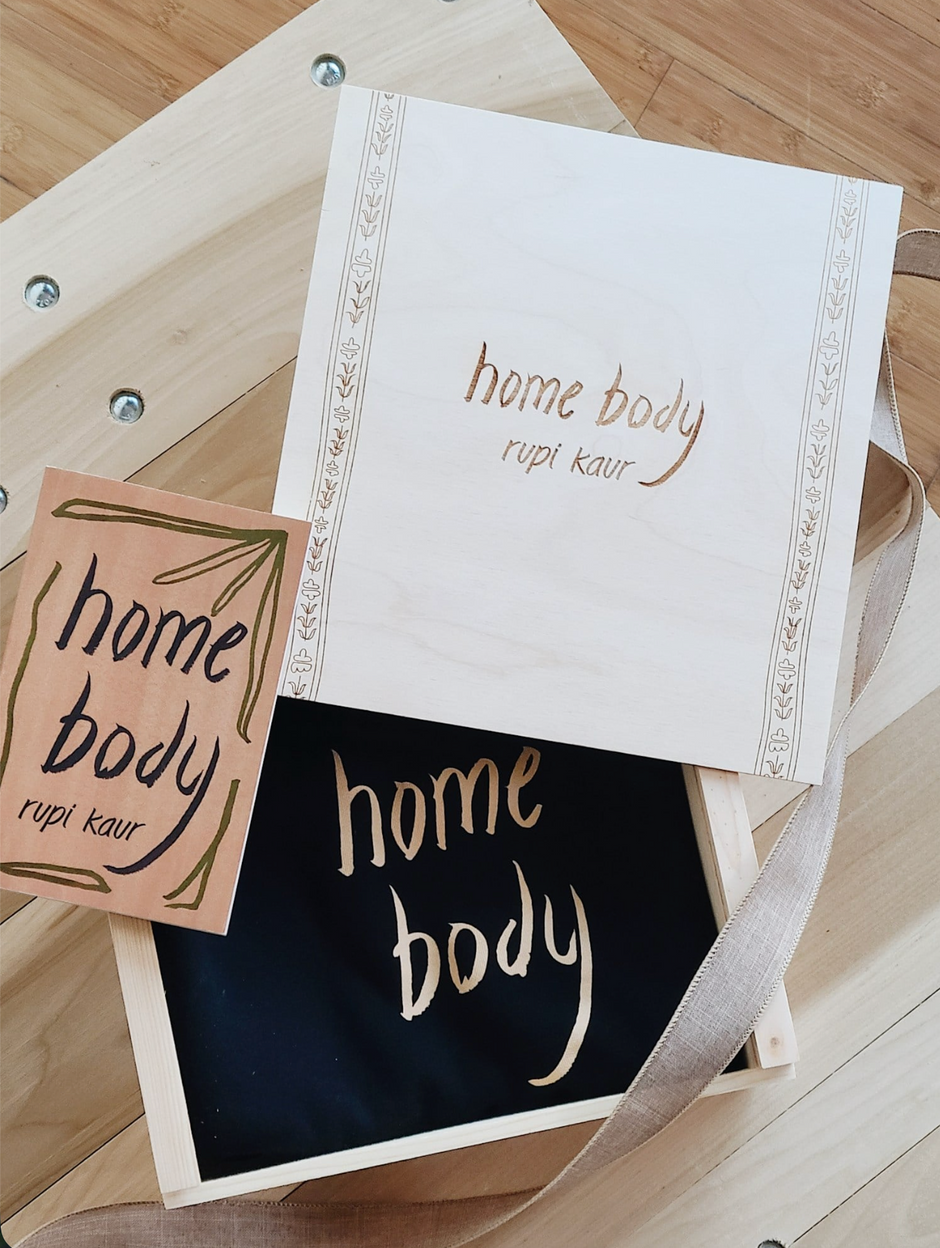 A custom wooden box lid, a book, and the inside of the custom wooden gift box, all with the words 'home body' and 'rupi kaur' arranged on a light wood surface. The book cover is cream with gold decorative border lines and gold lettering.