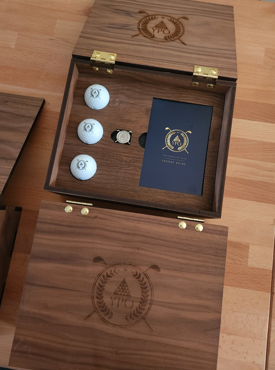 An open wooden presentation box on a wood floor, containing three white golf balls, a small round golf accessory, a dark blue card with gold lettering, and a matching wooden lid engraved with a golf-themed emblem.
