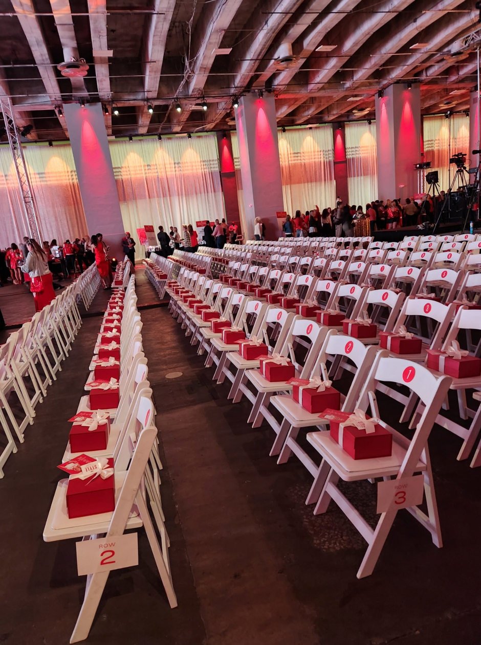 A large event hall with rows of white folding chairs arranged in aisles, each chair decorated with a red gift box and white bow. People are seated and standing in the background under warm red and white lighting.