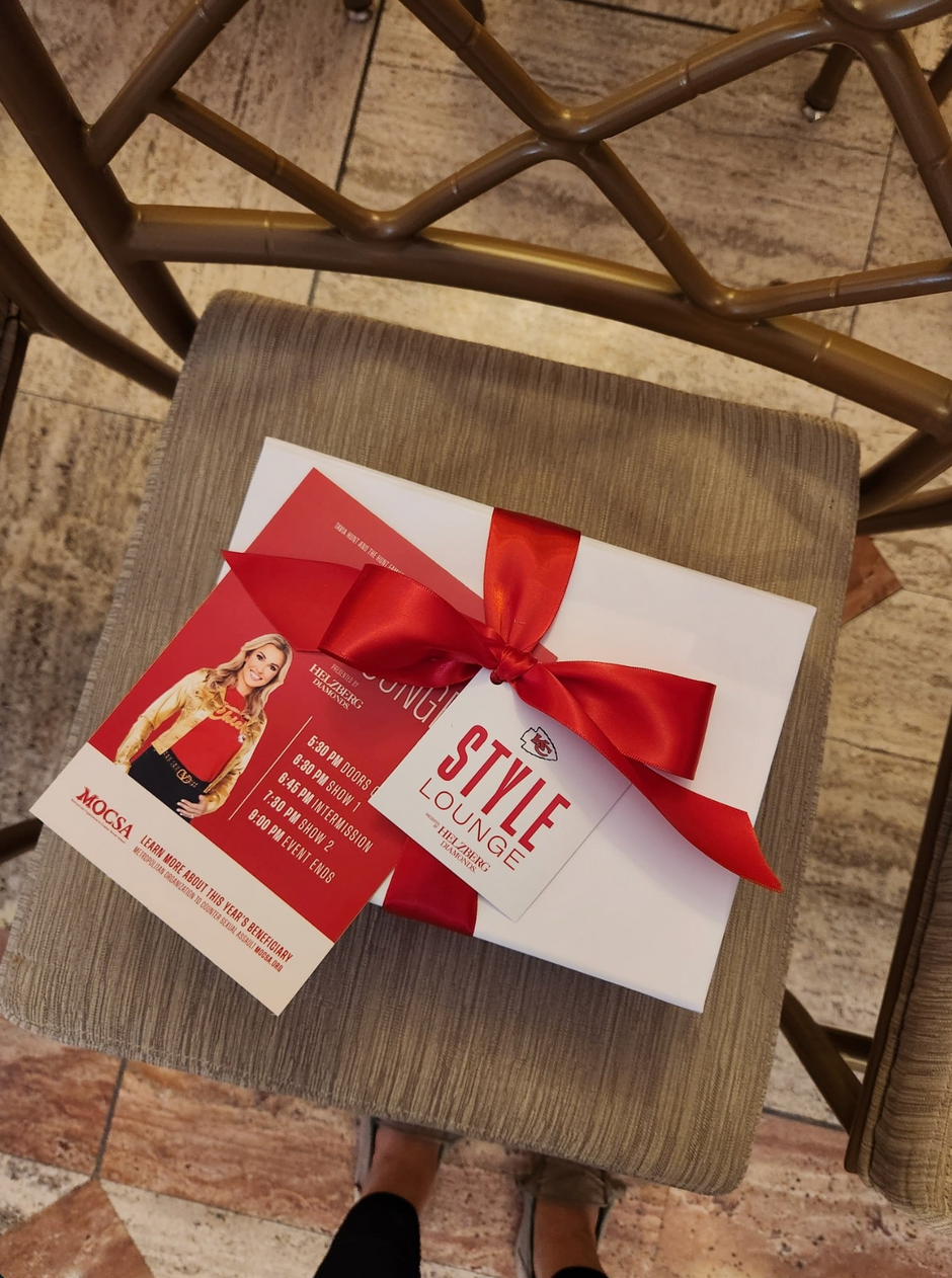 A white gift box tied with a red ribbon sits on a woven chair. Attached is a red and white card for MOCSA x Chiefs Style Lounge, featuring a photo of a woman and event text.