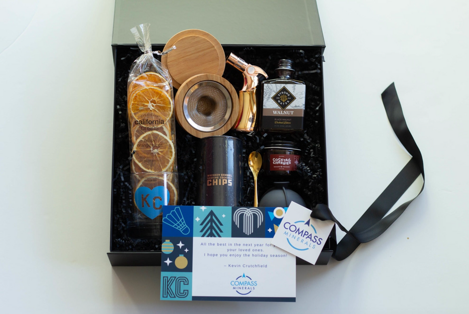 A curated gift box set displayed on a white surface, featuring a black box with black shredded filler containing a bag of dried orange slices, a bottle of walnut bitters, a cocktail smoker kit, a gold spoon, and cocktail cherries. A black ribbon is tied to the right side.