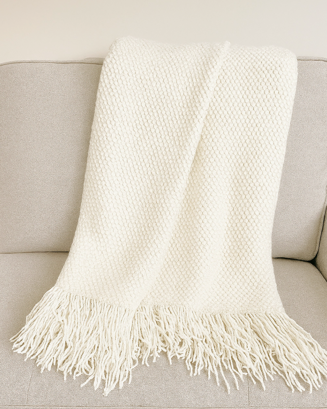 An off-white textured knit throw blanket with fringe, draped over a light beige sofa.