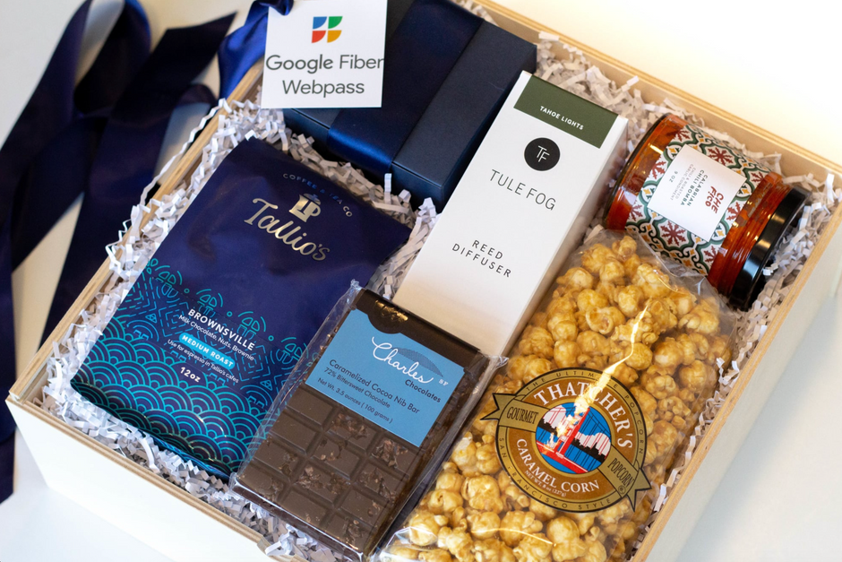 A curated gift box filled with assorted gourmet items, including a blue packet of Tallios Brownswille coffee, a bar of Charles chocolate, a box of Tule Fog reed diffuser, and a jar of Thatcher's Caramel Corn popcorn, arranged in white paper shred with a navy ribbon.
