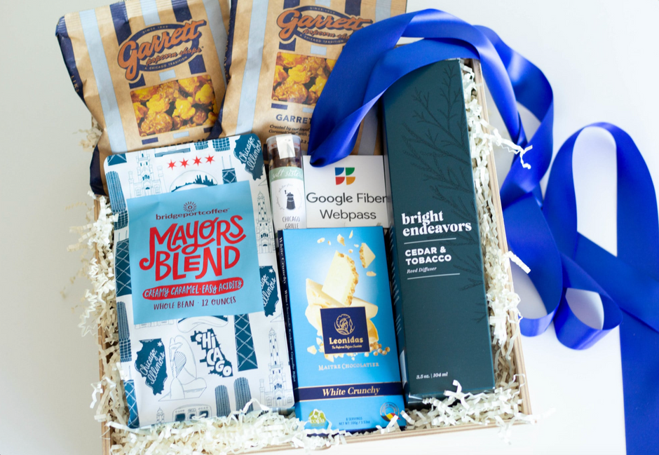 A curated gift box filled with assorted packaged snacks and beverages, including Garrett's Popcorn, Mayors Blend coffee, Google Fiber Webpass gift tag, and a dark box of cedar & tobacco scented reed diffuser, arranged in white shredded paper with a bright blue ribbon.