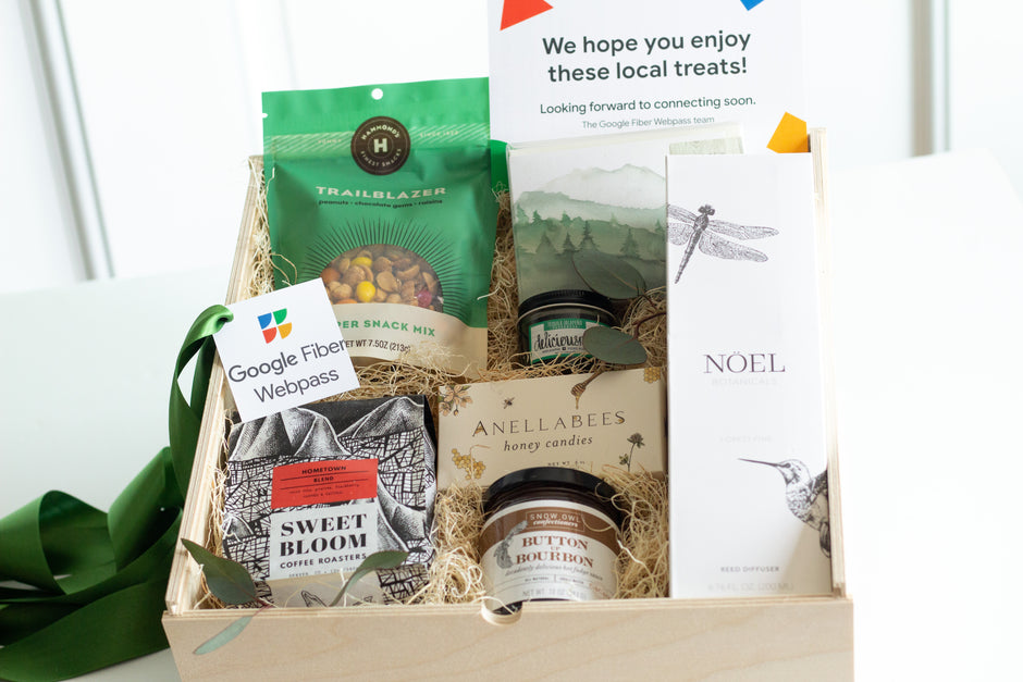 A curated gift box filled with assorted local treats and packaged snacks, including Trailblazer Trail Mix, Google Fiber Webpass tag, Sweet Bloom coffee, Anellabees honey candles, and a white box holding a reed diffuser from Noel, arranged in a light wood tray with shredded paper filler.