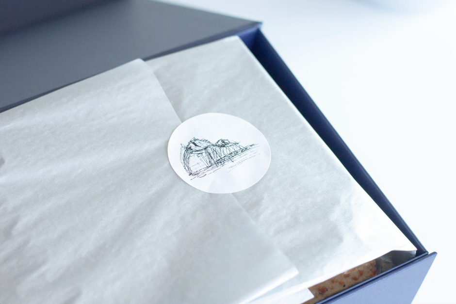A close-up of a dark blue gift box with white tissue paper inside and a round white sticker featuring a black line drawing of Kauffman Performing Arts Center. 