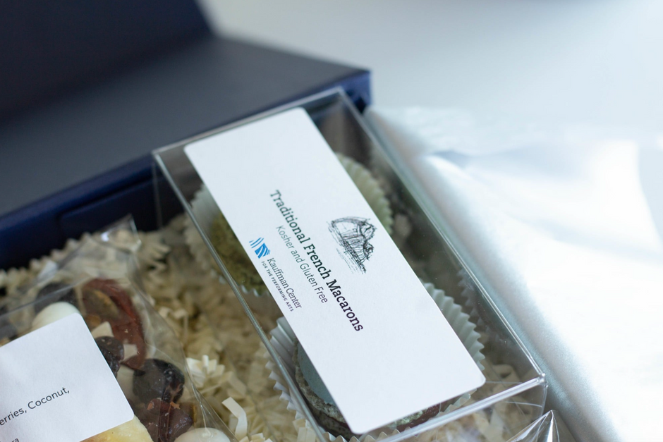 A clear plastic gift box filled with individually wrapped food items and shredded paper filler, with a white label reading 'Traditional French Macarons' and a small logo. Part of a navy blue box is visible behind it on a light surface.