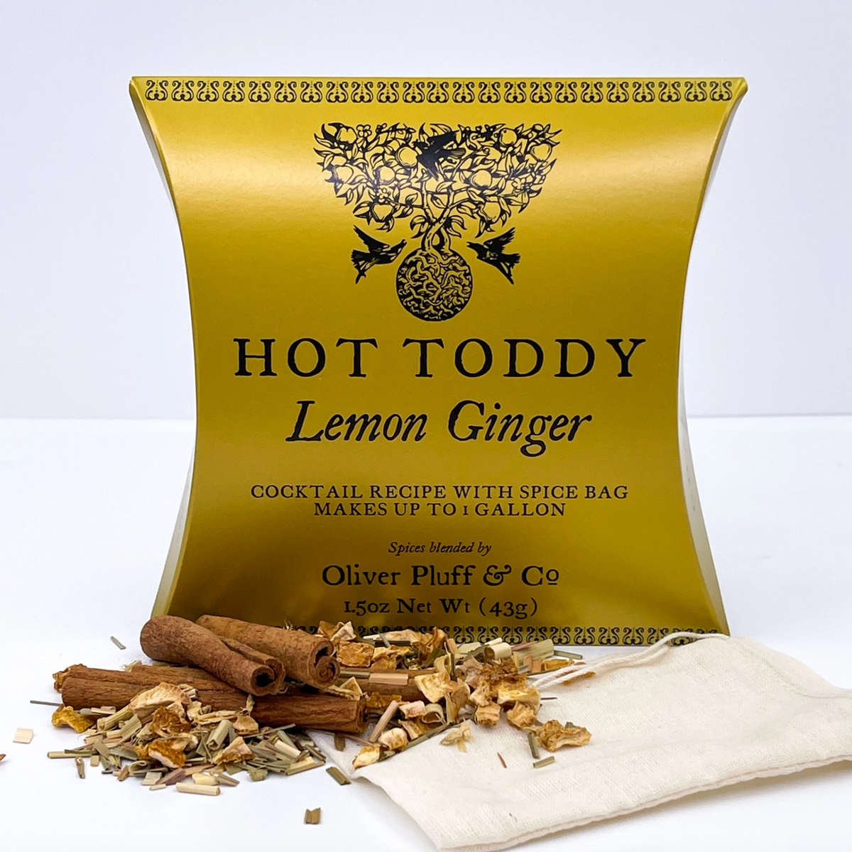 A gold metal tin labeled HOT TODDY Lemon Ginger stands on a white surface with loose spices and a small cloth bag in front. Visible text includes Oliver Pluff & Co.