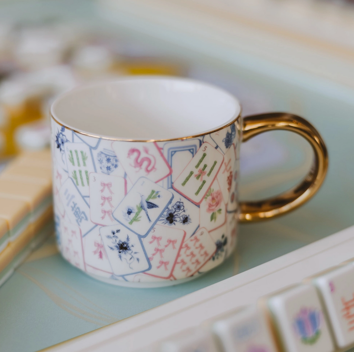 A white porcelain teacup decorated with a colorful mahjong tile pattern and featuring a shiny gold handle, resting on a light blue tray.