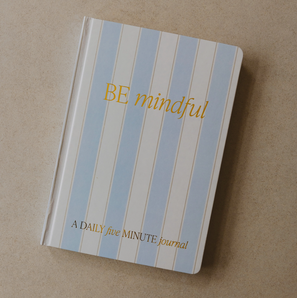 A daily five minute journal with a light blue and white striped cover, featuring gold foil text that reads 'BE mindful' and 'A DAILY five MINUTE Journal', photographed on a neutral beige background.
