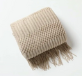 Load image into Gallery viewer, A folded chunky knit throw blanket in a light beige color, featuring a textured chevron pattern and fringed edges, photographed on a clean white background.
