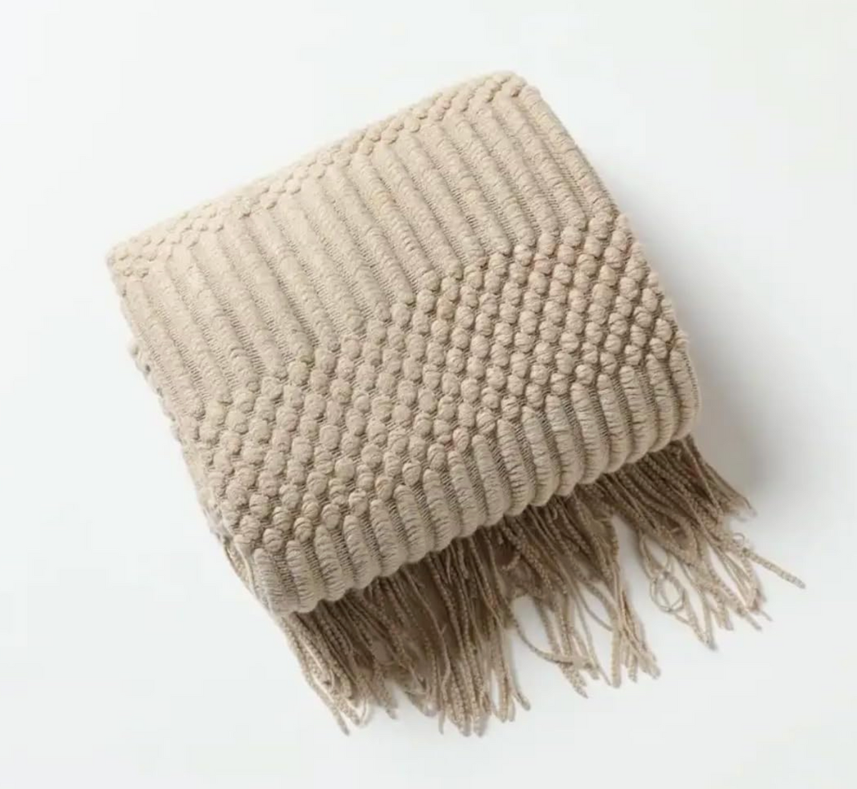 A folded chunky knit throw blanket in a light beige color, featuring a textured chevron pattern and fringed edges, photographed on a clean white background.
