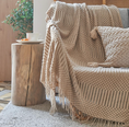 Load image into Gallery viewer, A cozy living room scene featuring a chunky knit throw blanket and matching pillow in a warm beige color draped over a sofa next to a rustic wooden side table.
