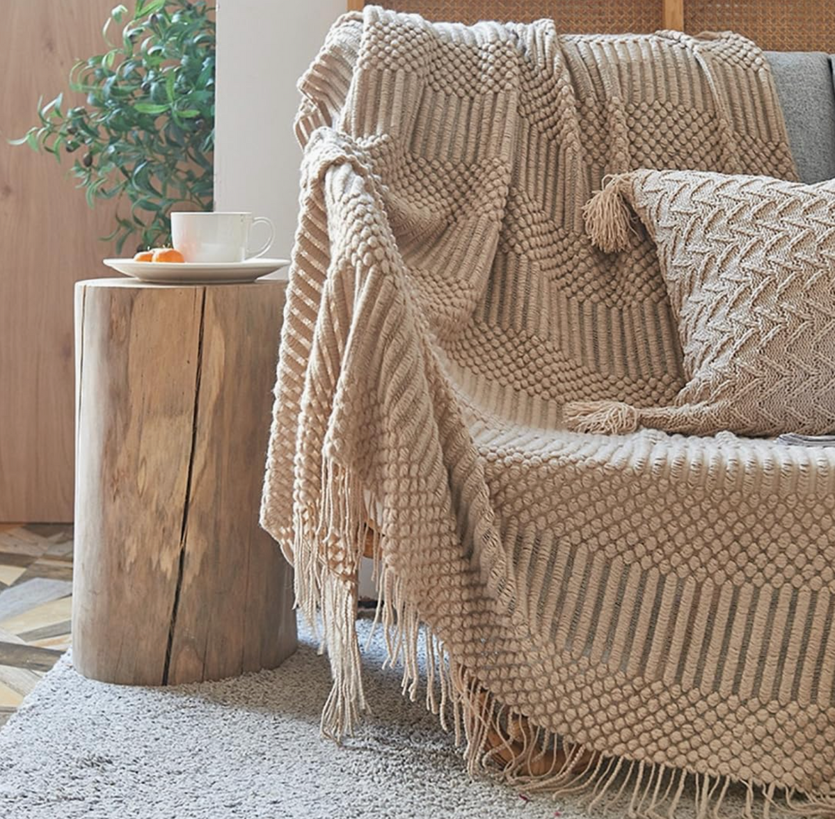A cozy living room scene featuring a chunky knit throw blanket and matching pillow in a warm beige color draped over a sofa next to a rustic wooden side table.