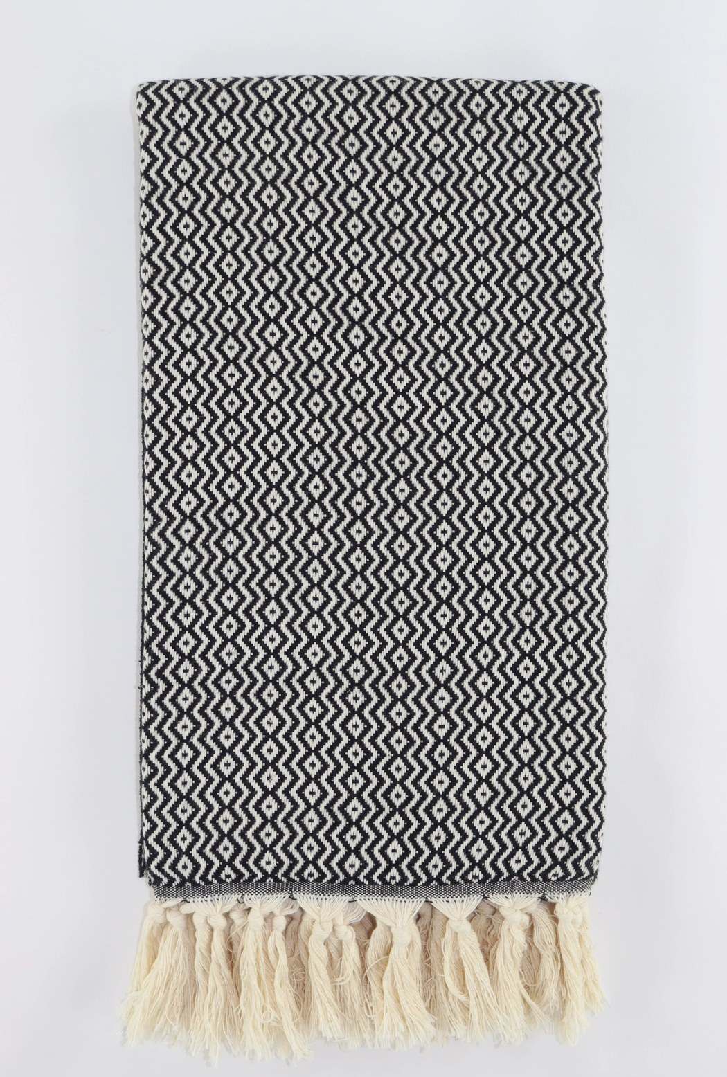 A folded woven turkish towel linen with a black and off-white geometric zigzag pattern and cream fringe tassels along one edge, shown against a plain light background.