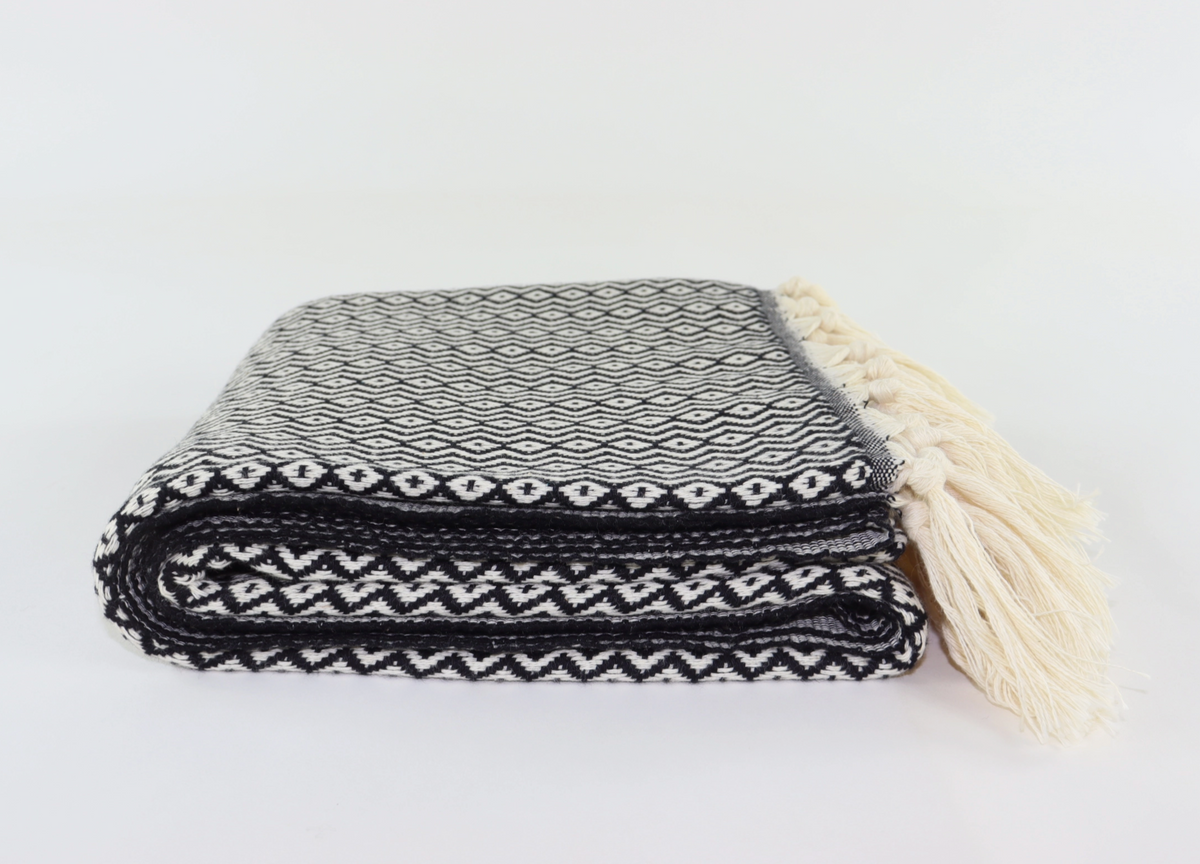 A folded black and white patterned woven turkish towel linen with cream-colored tassels on the side, set against a plain white background.