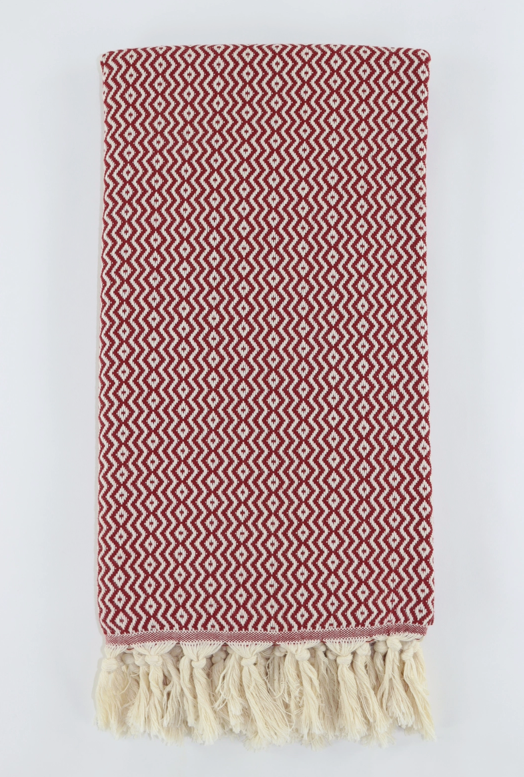 A folded woven turkish linen with a red and off-white geometric zigzag pattern and cream tassels along one edge, photographed against a light gray background.