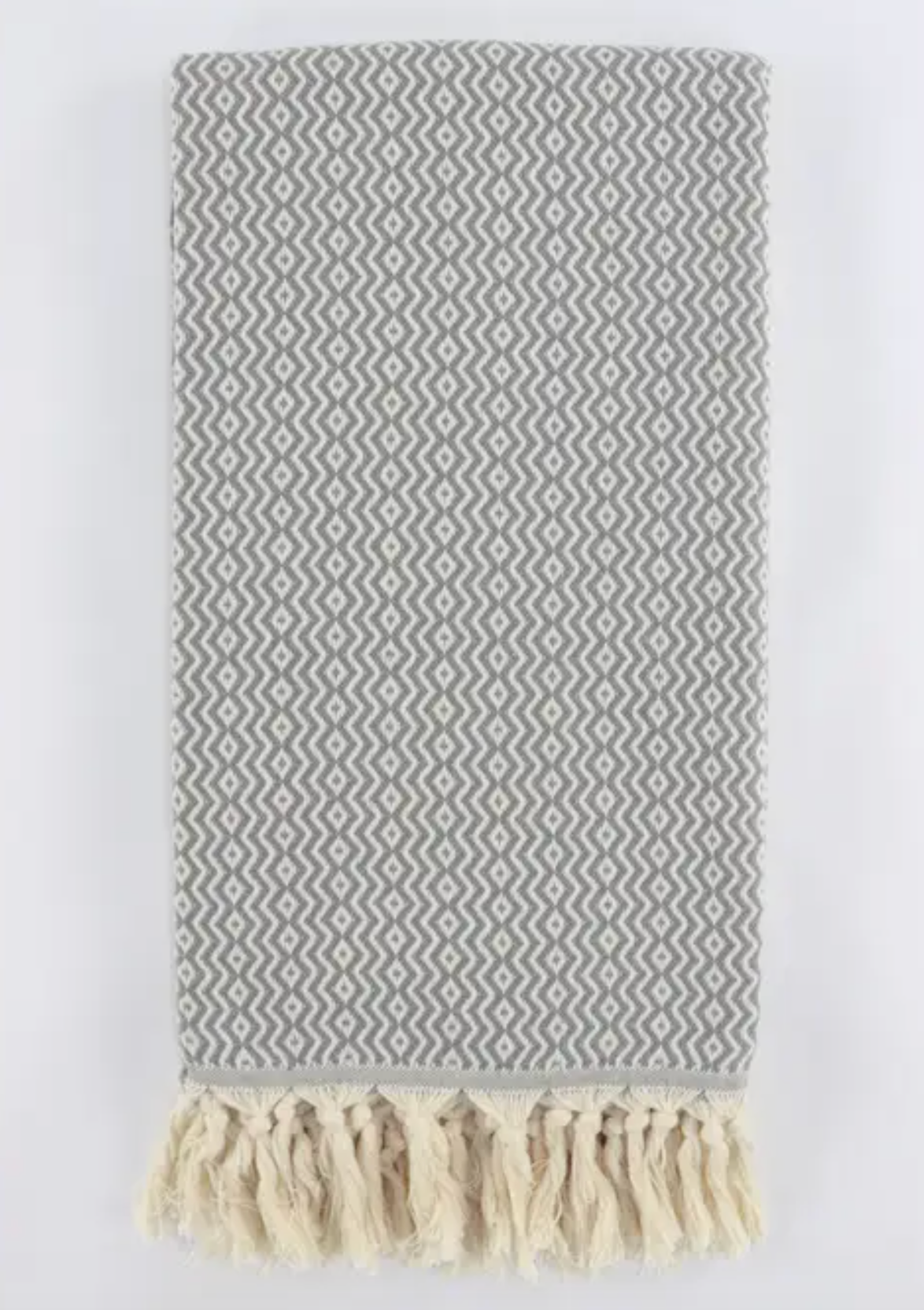 A folded woven turkish towel linen with a gray and off-white geometric diamond pattern and cream-colored fringe tassels at the bottom, shown against a plain white background.