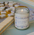 Load image into Gallery viewer, A lit candle in a clear glass jar sits on a tabletop with a blurred mahjong set in the background. The label reads 'Sweet Water Decor' and 'Playing Mahjong with Mah Girls'.
