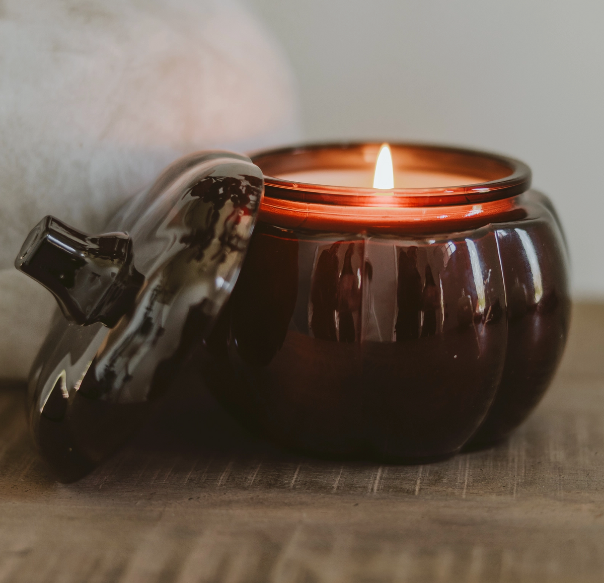 A dark amber glass candle jar shaped like a pumpkin, shown with its lid leaning against it on a wooden surface. The candle is lit, with a warm glow visible inside the jar.
