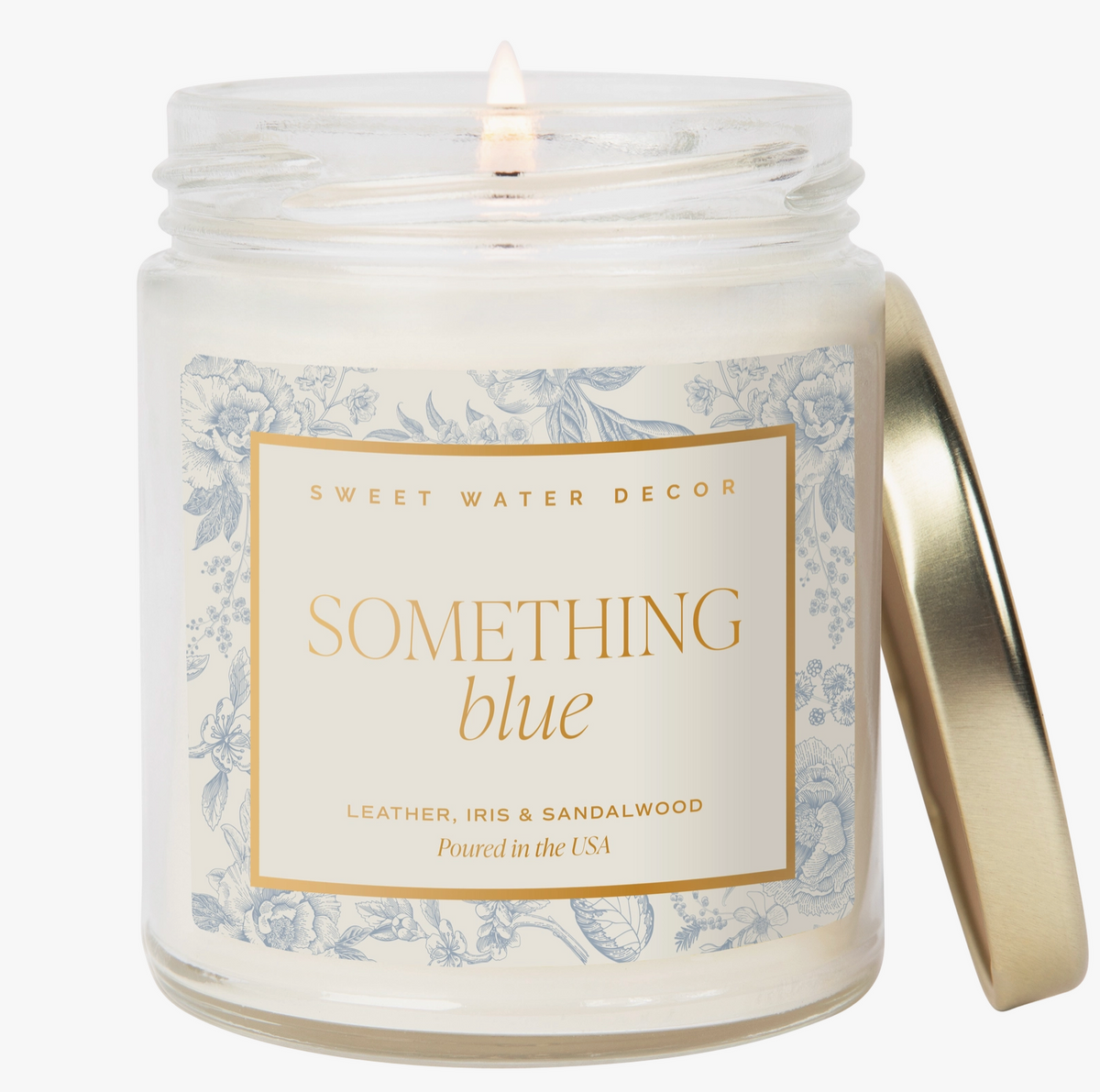 A glass jar candle with a white wax fill, a lit wick, a white label with blue floral border and gold text reading 'SOMETHING blue', and a metallic gold lid leaning against the right side on a white background.