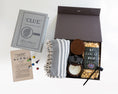 Load image into Gallery viewer, A flat-lay presentation of a 'Clue' themed mystery gift box set on a white background. The set includes a vintage-style clue book, a printed clue card, several colored play pieces, a gray and white patterned throw blanket, a dark leather set of coasters, a pumpkin candle, a bag of KC Local Pop popcorn, and a small black candle snuffer inside an open dark brown box.
