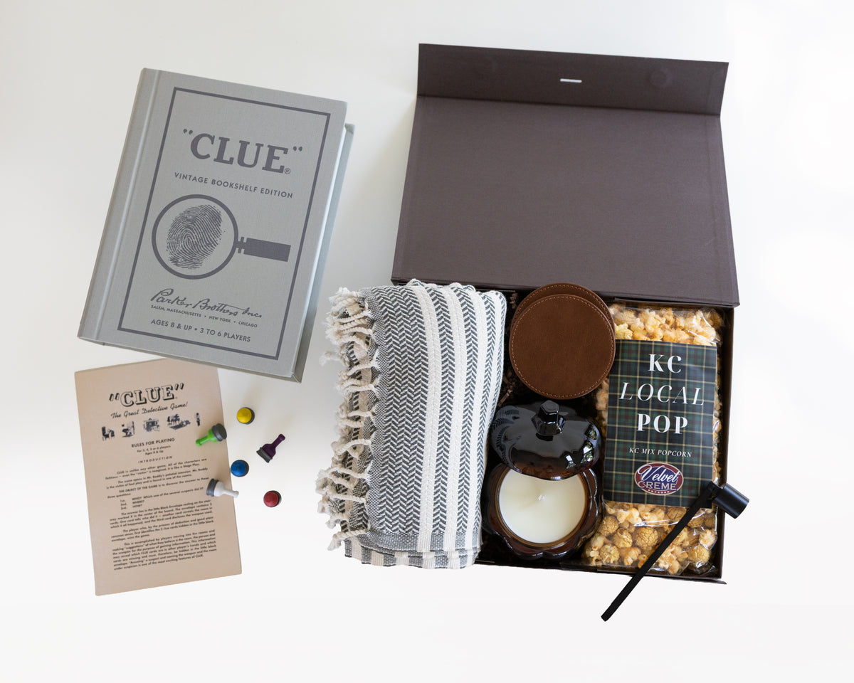 A flat-lay presentation of a 'Clue' themed mystery gift box set on a white background. The set includes a vintage-style clue book, a printed clue card, several colored play pieces, a gray and white patterned throw blanket, a dark leather set of coasters, a pumpkin candle, a bag of KC Local Pop popcorn, and a small black candle snuffer inside an open dark brown box.