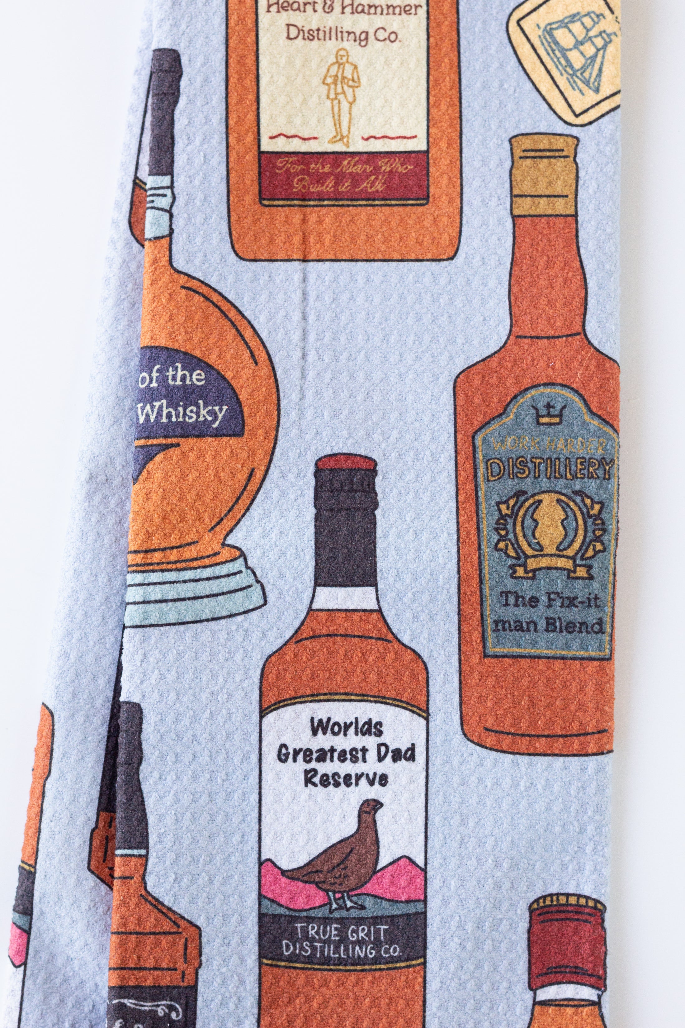 A light blue kitchen towel featuring a repeating pattern of illustrated whiskey and spirit bottles in orange, brown, black, and cream, with humorous labels like 'World's Greatest Dad Reserve' and 'True Grit Distilling Co.'.